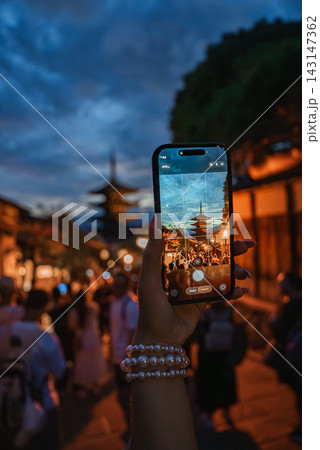 A hand holds a smartphone framing Yasaka Pagoda in Kyoto at dusk, lantern lit machiya line Ninenzaka and Sannenzaka, blurred crowd moves, warm lights and blue clouds set mood A hand holds a smartphone framing Yasaka Pagoda in Kyoto at dusk, lantern lit machiya line Ninenzaka and Sannenzaka, blurred crowd moves, warm lights and blue clouds set mood 143147362