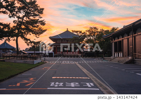 A wide road leads to a wooden temple hall in Nara, Japan, at dusk. Tall pines line the way, long corridors flank the path, and warm light casts soft shadows. A wide road leads to a wooden temple hall in Nara, Japan, at dusk. Tall pines line the way, long corridors flank the path, and warm light casts soft shadows. 143147424