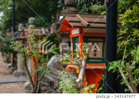 Traditional stone lanterns and orange and green lamps line a gravel path in Kyoto, with kanji signage visible. Soft overcast light and shallow focus highlight wood and stone. Traditional stone lanterns and orange and green lamps line a gravel path in Kyoto, with kanji signage visible. Soft overcast light and shallow focus highlight wood and stone. 143147477