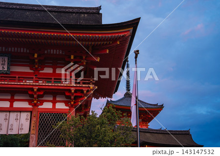 A vermilion temple gate and tiered pagoda stand at Kiyomizu dera in Kyoto at dusk. Warm lights reveal eaves, latticework, and a Japanese flag in soft blue light. 143147532