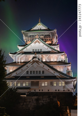 Osaka Castle stands lit at night in Osaka, Japan, with green and purple aurora like bands above. The centered upward view shows layered walls, roofs, and gold details. 143147773