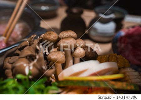 Clusters of shimeji at center with maitake, kamaboko, corn, greens, and thin meats, chopsticks and pot blur behind. Warm low light and shallow depth of field in Osaka. 143147806