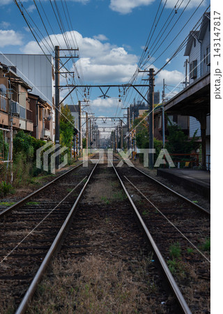Symmetrical tracks run through a quiet Osaka neighborhood, with wooden houses, small platforms, catenary wires, and poles guiding the gaze to a distant vanishing point. 143147817
