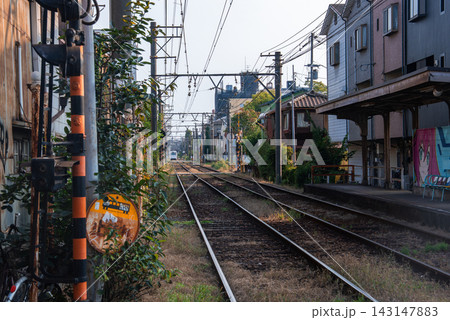 Twin tracks pass low rise houses in Osaka, Japan, with a modest platform, convex mirror, striped crossing pole, and gantries in warm late afternoon light. 143147883