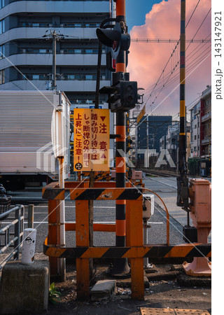 Street level view in Osaka shows a railway crossing with black and orange barriers, signal lights, and a truck passing, late afternoon light adds a warm cinematic mood. 143147921