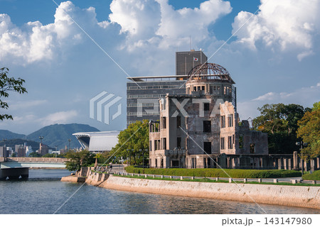 The Hiroshima Peace Memorial stands by the Motoyasu River in Hiroshima, Japan. People stroll along the promenade as modern buildings and distant hills frame the ruin. The Hiroshima Peace Memorial stands by the Motoyasu River in Hiroshima, Japan. People stroll along the promenade as modern buildings and distant hills frame the ruin. 143147980