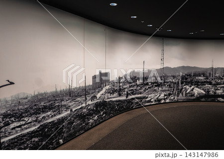 A panoramic black and white mural wraps a museum gallery in Hiroshima, showing ruined streets, skeletal trees, and a lone mid rise building as visitors walk along. A panoramic black and white mural wraps a museum gallery in Hiroshima, showing ruined streets, skeletal trees, and a lone mid rise building as visitors walk along. 143147986