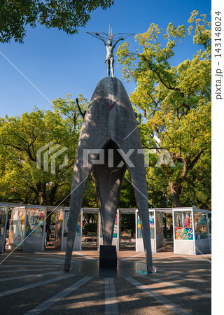 The monument in Hiroshima Peace Memorial Park shows the girl with a crane, arching concrete form, and display cases of origami cranes and artwork under green trees. The monument in Hiroshima Peace Memorial Park shows the girl with a crane, arching concrete form, and display cases of origami cranes and artwork under green trees. 143148024