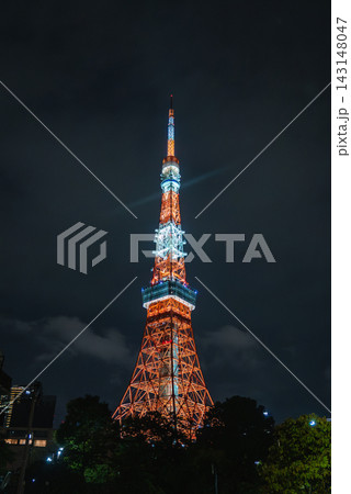 Tokyo Tower stands lit in Minato, Tokyo at night, cool white bands near decks, treetops and low buildings frame the base, centered composition highlights design Tokyo Tower stands lit in Minato, Tokyo at night, cool white bands near decks, treetops and low buildings frame the base, centered composition highlights design 143148047