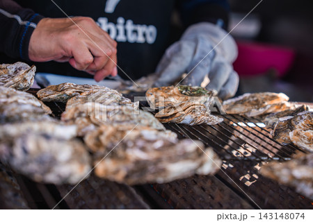 Two people grill and shuck large oysters on a metal grate on Itsukushima near Hiroshima, Japan. Close hands, one gloved, work rows of rough shells in tight focus. Two people grill and shuck large oysters on a metal grate on Itsukushima near Hiroshima, Japan. Close hands, one gloved, work rows of rough shells in tight focus. 143148074