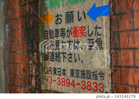 A worn Japanese notice sign on a brick wall shows multicolored text, arrows, and a bold red emergency number, suggesting a railway underpass in Tokyo. 143148179