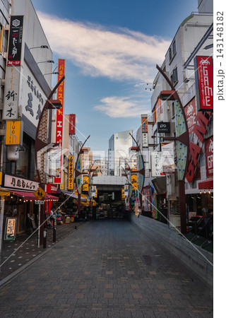 A quiet pedestrian alley in Tokyo near Ameya Yokocho shows shops, arcades, and an overhead rail bridge, with GAME Taito Station sign and yellow banners in early light. 143148182