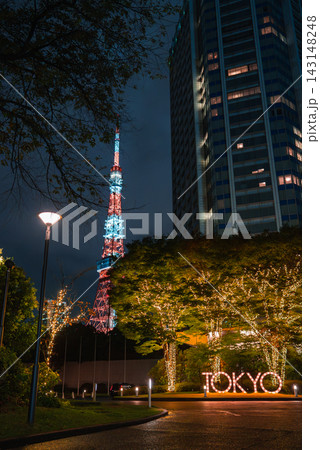 Night scene in Tokyo, with Tokyo Tower behind a high rise, trees wrapped in warm lights along a walkway, and a glowing TOKYO sign at a plaza in Minato. 143148248