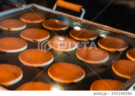 Golden brown dorayaki pancakes puff as they cook on a large griddle at a Tokyo street market, even rows on a shiny metal surface suggest a vendor setup. Golden brown dorayaki pancakes puff as they cook on a large griddle at a Tokyo street market, even rows on a shiny metal surface suggest a vendor setup. 143148290
