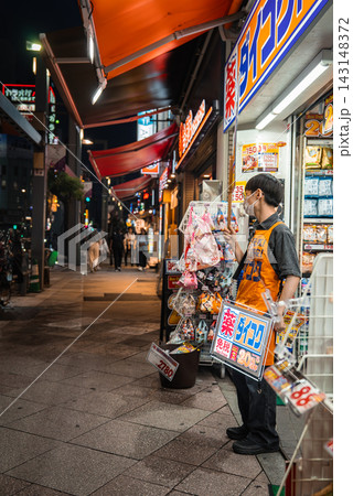 A vendor in an orange apron holds signs outside a Tokyo drugstore as evening shoppers pass in a neon lit district with red awnings and busy retail displays. A vendor in an orange apron holds signs outside a Tokyo drugstore as evening shoppers pass in a neon lit district with red awnings and busy retail displays. 143148372