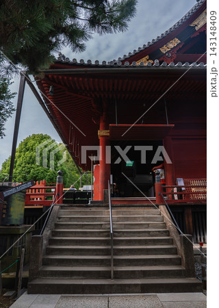 Stone steps rise to a deep red wooden hall at Senso ji in Asakusa, Tokyo, with lanterns, lattice details, eaves, and crimson pillars under a green tree canopy. 143148409