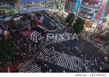 Hundreds cross the diagonal crosswalks at Shibuya Crossing in Tokyo, umbrellas moving past neon storefronts and billboards as traffic lines buses by Shibuya Station. Hundreds cross the diagonal crosswalks at Shibuya Crossing in Tokyo, umbrellas moving past neon storefronts and billboards as traffic lines buses by Shibuya Station. 143148422