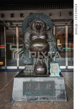 A bronze tanuki stands at a Tokyo station entrance, straw hat on, sake bottle and ledger in hand, round belly forward, plaque at its feet, travelers moving past. 143148424