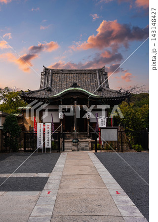 A small Shinto shrine in Tokyo at dusk stands at a stone path end, with a curved tiled roof, hanging lanterns, rope with paper shide, banners, and a low gate among trees. A small Shinto shrine in Tokyo at dusk stands at a stone path end, with a curved tiled roof, hanging lanterns, rope with paper shide, banners, and a low gate among trees. 143148427