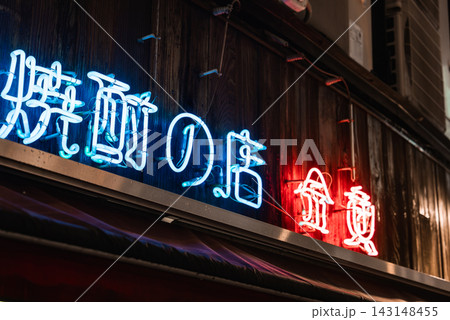 Close view of a wooden storefront in Tokyo shows blue and red neon Japanese characters, angled composition, night lighting, hinting at a shochu or izakaya bar. Close view of a wooden storefront in Tokyo shows blue and red neon Japanese characters, angled composition, night lighting, hinting at a shochu or izakaya bar. 143148455