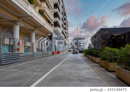 Soft light reflects on Monaco harbor balconies and a lattice pavilion as cars drive past crash barriers and red bollards toward Port Hercules with yachts and masts. Soft light reflects on Monaco harbor balconies and a lattice pavilion as cars drive past crash barriers and red bollards toward Port Hercules with yachts and masts. 143148473