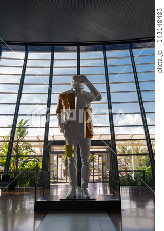 A white astronaut statue with gold fabric and bag stands in a glass atrium in Nice, France. Tall slatted windows reveal sky and palm trees, shot low with soft daylight. A white astronaut statue with gold fabric and bag stands in a glass atrium in Nice, France. Tall slatted windows reveal sky and palm trees, shot low with soft daylight. 143148483