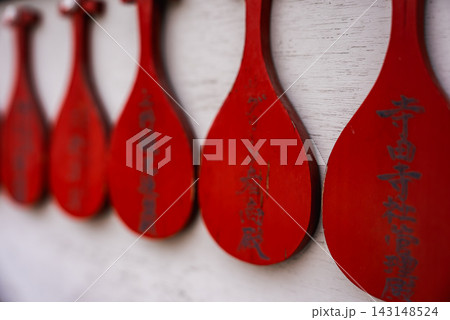 A row of red wooden rice paddles with vertical Japanese text lines a pale wall at a Tokyo shrine, shallow depth of field draws focus to carved characters in front. 143148524