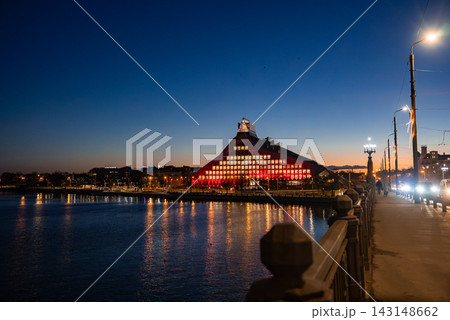 Twilight view in Riga, Latvia shows the National Library of Latvia lit red, windows glowing and reflecting on the Daugava, seen from a bridge with lamps and traffic during Staro Riga 2025. Twilight view in Riga, Latvia shows the National Library of Latvia lit red, windows glowing and reflecting on the Daugava, seen from a bridge with lamps and traffic during Staro Riga 2025. 143148662
