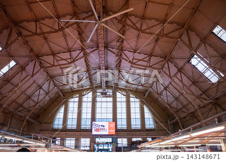 Daytime view inside Riga Central Market in Latvia shows arched windows, steel trusses, and a large ceiling fan above vendor stalls with light on a digital sign. 143148671