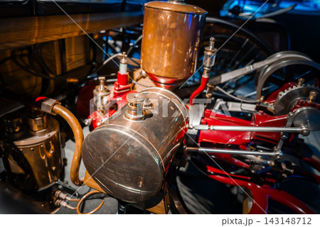 Close up of polished brass and copper engine parts with red linkages in an auto museum, cool toned lighting, detailing seams, reservoir, and exposed rods. 143148712