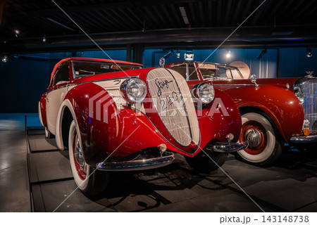 A red and cream vintage coupe stands under museum lighting, with sweeping fenders, teardrop headlights, and a tall chromed grille, classic cars lined on polished floors. 143148738