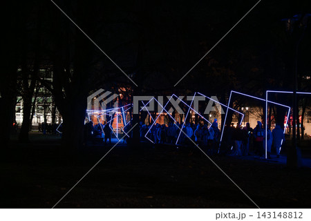 Visitors walk under tilted rectangles and diamonds in a tree lined park in central Riga, Latvia. Blue LED frames form a glowing corridor as city buildings glow softly. Visitors walk under tilted rectangles and diamonds in a tree lined park in central Riga, Latvia. Blue LED frames form a glowing corridor as city buildings glow softly. 143148812
