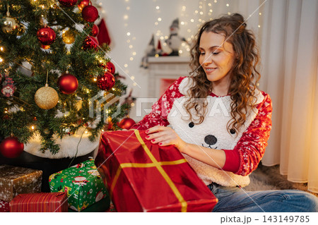 Smiling woman opening a red Christmas gift by decorated tree Smiling woman opening a red Christmas gift by decorated tree 143149785