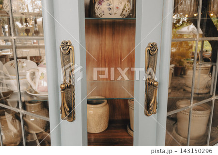 Vintage wooden cabinet with brass handles and glass doors storing ceramic dishware Vintage wooden cabinet with brass handles and glass doors storing ceramic dishware 143150296