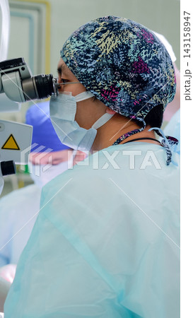 Focused surgeon wearing patterned cap, mask, protective eyewear, and sterile gown while using operating microscope during delicate procedure inside clinical surgical room Focused surgeon wearing patterned cap, mask, protective eyewear, and sterile gown while using operating microscope during delicate procedure inside clinical surgical room 143158947