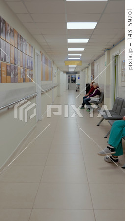 Hospital corridor with seated patients waiting on chairs beneath fluorescent ceiling lights, handrails, wall posters, glossy tiled floor, calm clinical interior perspective Hospital corridor with seated patients waiting on chairs beneath fluorescent ceiling lights, handrails, wall posters, glossy tiled floor, calm clinical interior perspective 143159201