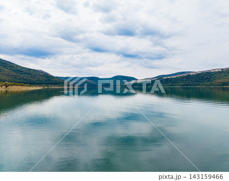 View of a calm lake surrounded by mountains on a cloudy day with reflections on the water View of a calm lake surrounded by mountains on a cloudy day with reflections on the water 143159466