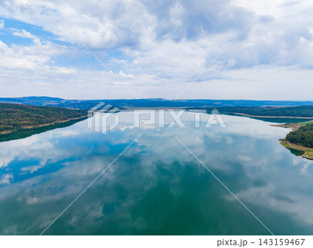 Calm water reflects clouds above in a wide lake near green hills during a sunny day Calm water reflects clouds above in a wide lake near green hills during a sunny day 143159467
