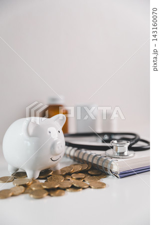 White piggy bank surrounded by coins, stethoscope, notebook and medical supplies, representing healthcare costs, medical savings, treatment expenses and financial planning for health emergencies. White piggy bank surrounded by coins, stethoscope, notebook and medical supplies, representing healthcare costs, medical savings, treatment expenses and financial planning for health emergencies. 143160070
