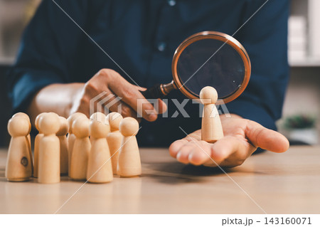 Magnifying glass evaluates a wooden candidate presented on an open hand beside a group, symbolizing recruitment, employee screening, talent search, assessment, hiring, selection, and human resources. Magnifying glass evaluates a wooden candidate presented on an open hand beside a group, symbolizing recruitment, employee screening, talent search, assessment, hiring, selection, and human resources. 143160071