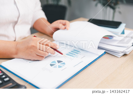 Business professional reviewing financial reports with a pen and turning document pages at an office desk, representing accounting, audit, data analysis, performance review, planning, Business professional reviewing financial reports with a pen and turning document pages at an office desk, representing accounting, audit, data analysis, performance review, planning, 143162417
