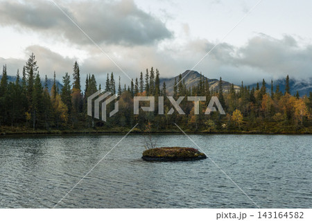 Small grassy island in a northern mountain lake. Beautiful scenic autumn landscape with a calm forest pond, spruce trees, and misty mountain peak under a cloudy sky. Small grassy island in a northern mountain lake. Beautiful scenic autumn landscape with a calm forest pond, spruce trees, and misty mountain peak under a cloudy sky. 143164582