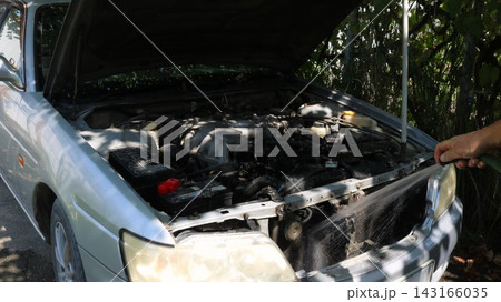 The process of washing a car's engine bay. A silver car is parked with its hood open, and a person is directing a jet of water from a hose onto the radiator. The process of washing a car's engine bay. A silver car is parked with its hood open, and a person is directing a jet of water from a hose onto the radiator. 143166035