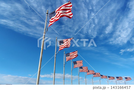 A row of American flags waving in the wind against a blue sky. A low-angle view of US flags on tall flagpoles and white clouds on a sunny day. Patriotic background. 143166457