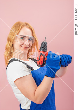 Confident young woman with red hair wearing protective goggles and work gloves, holding a cordless power drill, ready for home improvement or construction tasks against a pink background 143168599