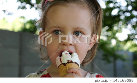 Portrait of attractive small girl eating delicious ice cream in a waffle cone outdoor. Beautiful child biting cold sweet dessert in summer garden. Cute kid enjoying tasty refreshing food on sunny day 143169089