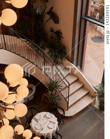modern, elegant staircase in an interior space with tables and chairs, plants, and a light fixture, viewed from an aerial perspective, featuring beige and black tones. modern, elegant staircase in an interior space with tables and chairs, plants, and a light fixture, viewed from an aerial perspective, featuring beige and black tones. 143169591