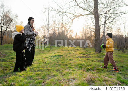 Parent and two children playing catch with ball in autumn park at sunset, low angle view with sun flare through leafless branches and warm backlight, playful moment together 143171426