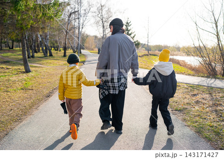 Mother walking with two children hand in hand along park path in autumn sunlight, rear view showing warm jackets, knit hats, and calm family outdoor bonding Mother walking with two children hand in hand along park path in autumn sunlight, rear view showing warm jackets, knit hats, and calm family outdoor bonding 143171427