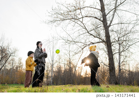 Parent and two children playing catch with ball in autumn park at sunset, low angle view with sun flare through leafless branches and warm backlight, playful moment together 143171441